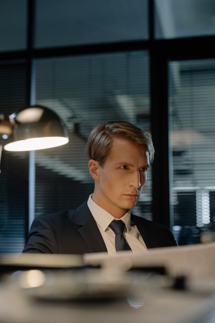 Serious detective in a suit examining documents under a desk lamp at night in a modern office.